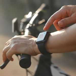 Female cyclist hands with heart rate icon on smart watch, bicycle in summer trail. All on screen are designed up.