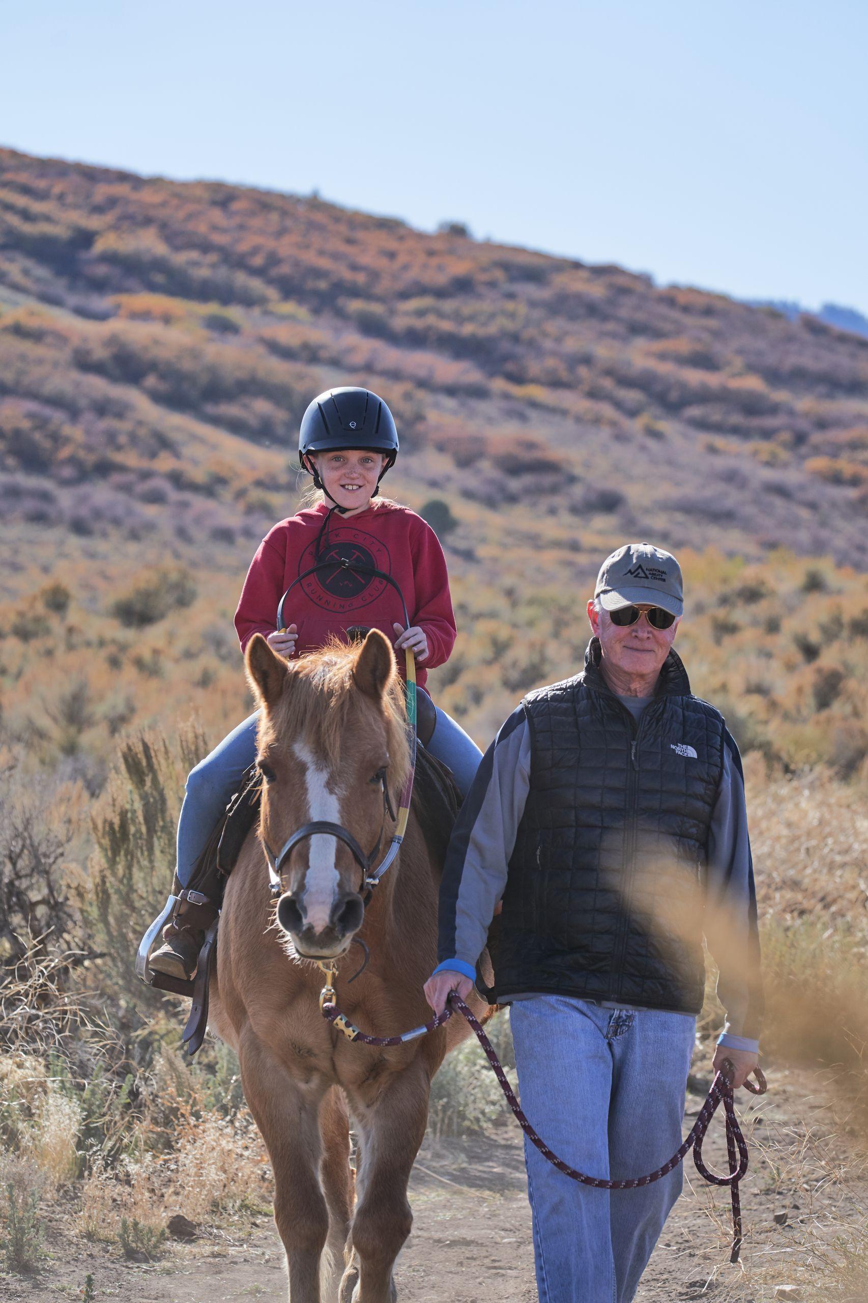 A young girl riding a horse is lead by a National Ability Center volunteer during an adaptive horseback riding lesson.