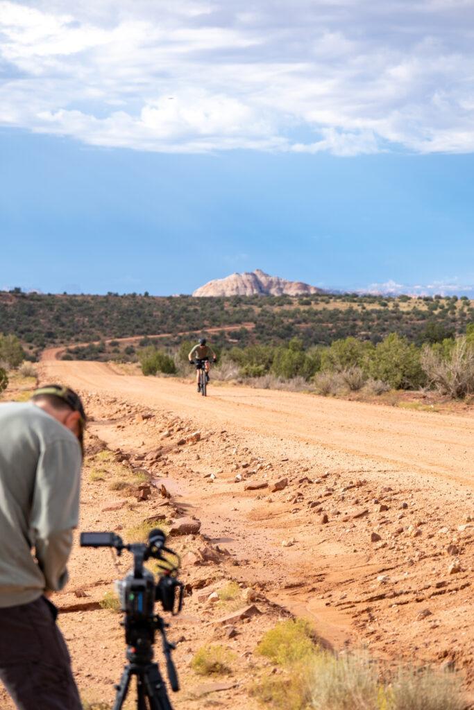 A cameraman in the foreground sets up a shot of a cyclist coming towards him. 