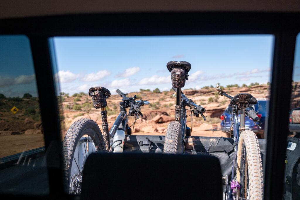 Three bikes in the back of a truck as seen through the rear window. 