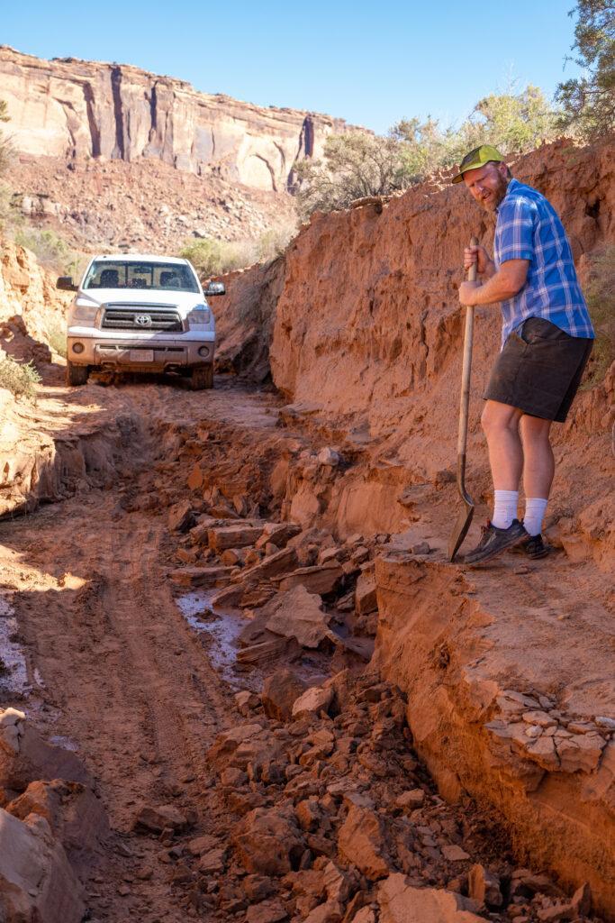 A person uses a shovel to level off a section of dirt road for a truck to pass. 