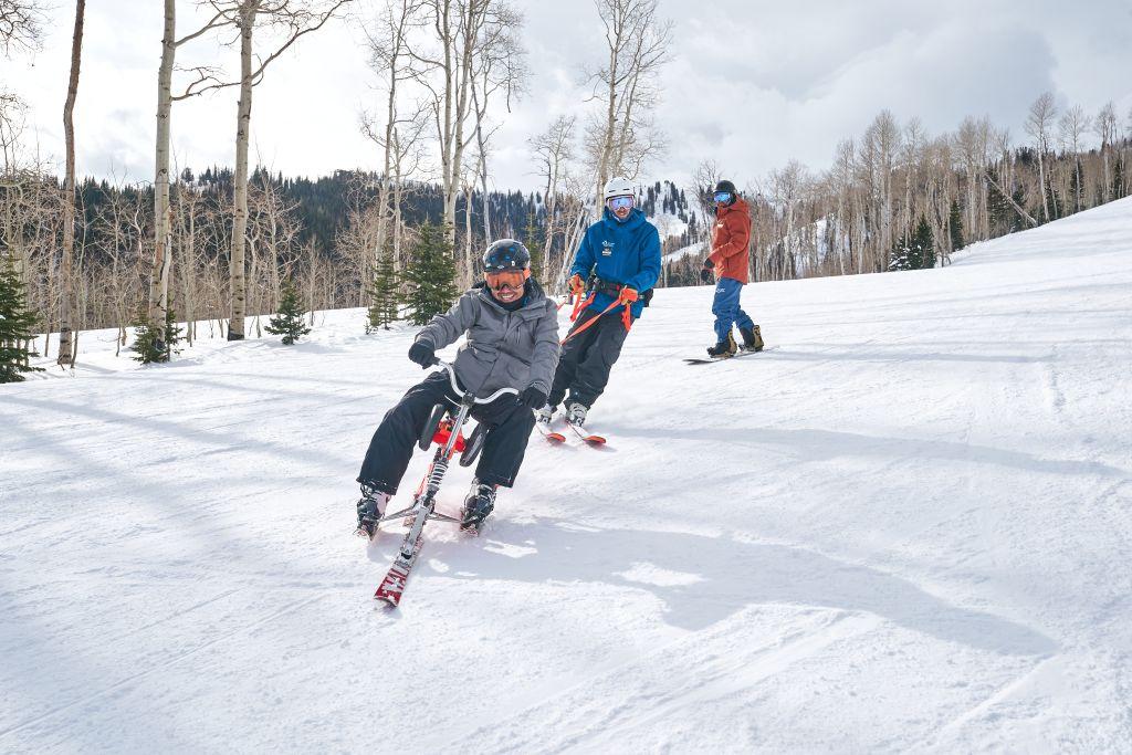 National Ability Center Participant slides on snow using a ski bike with an instructor tethering from behind. 