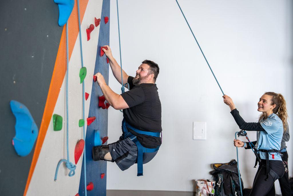 A double above the knee amputee climbs a wall at the National Ability Center using a seated harness and a belayer. 