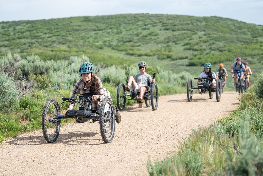 Multiple adaptive mountain bikers ride specialized adaptive bikes on a trail in Park City, Utah. 